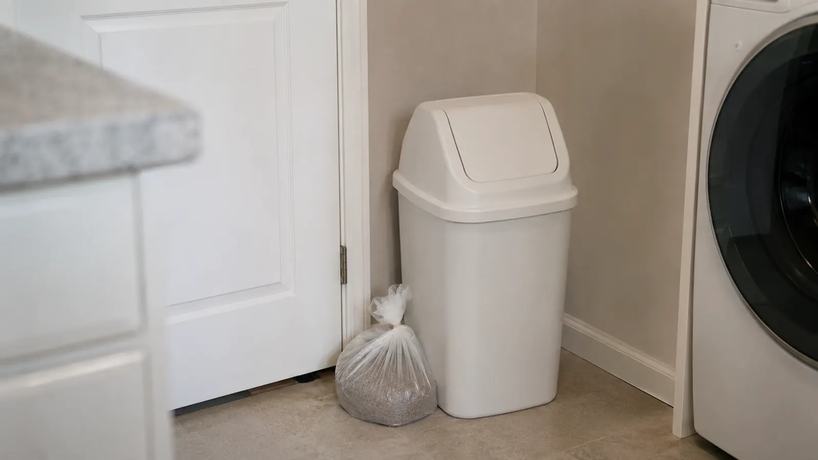 An ordinary kitchen trash can with a lid in the corner of a laundry room, a tied litter bag leaning beside it